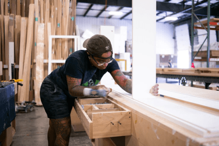 Craftsperson at Murphy Door® workshop carefully assembling a wooden hidden door drawer frame in a warehouse with lumber storage.
