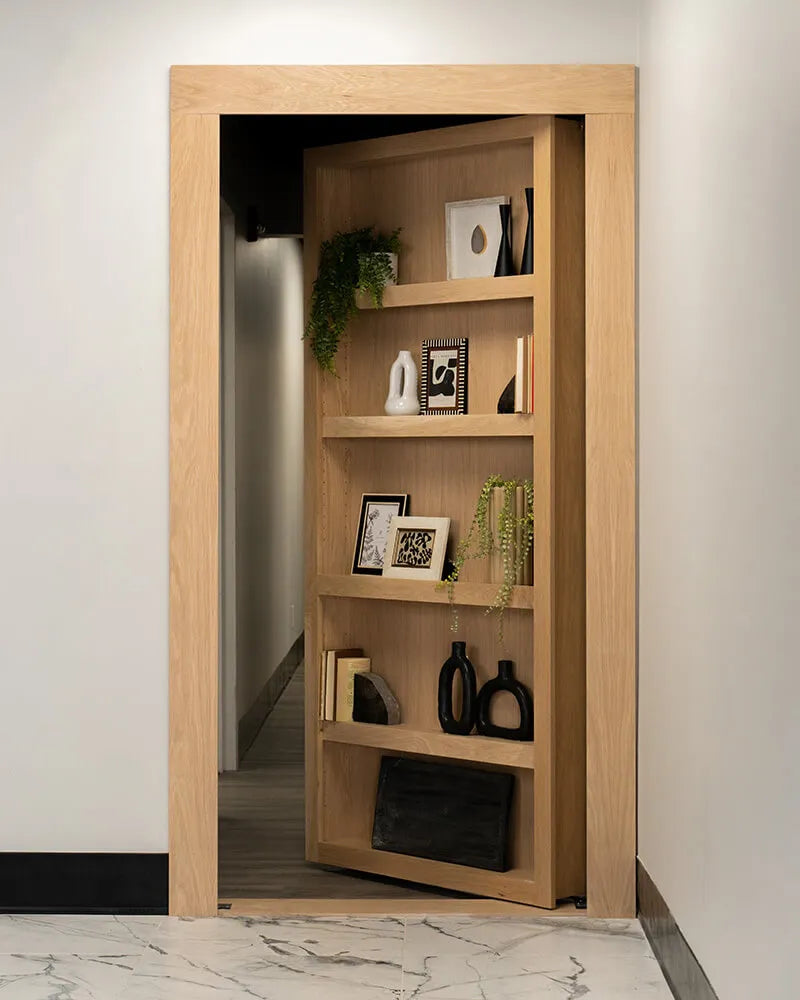 Murphy Door® wooden bookcase hidden door in light oak with decorative plants, frames, and vases on shelves, revealing hallway passage.