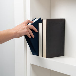 Hand reaching for navy blue book on white shelf containing Murphy Door® hidden door books in blue and black covers.