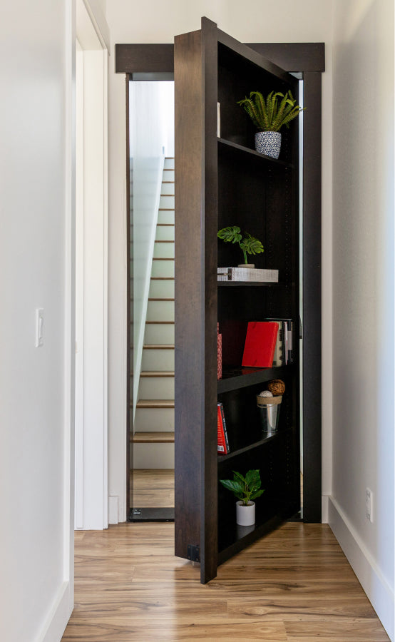 Murphy Door® dark wood bookcase hidden door partially open revealing staircase, with plants and books on shelves.