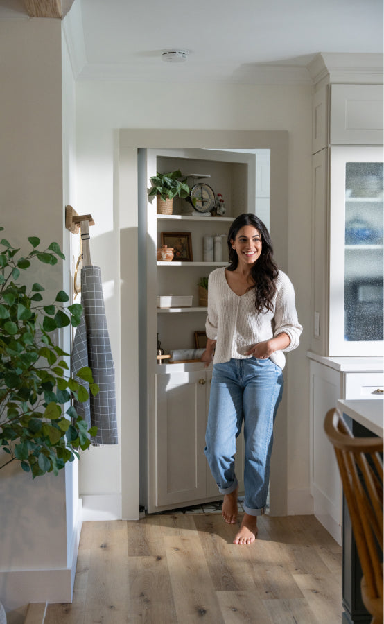 Woman in white sweater and jeans by Murphy Door® hidden pantry with shelving and decorative items in bright modern kitchen.