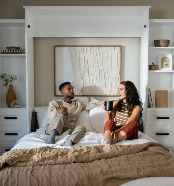 Two people enjoying coffee in a Murphy Door® wall bed with built-in white shelving and storage in a minimalist bedroom.