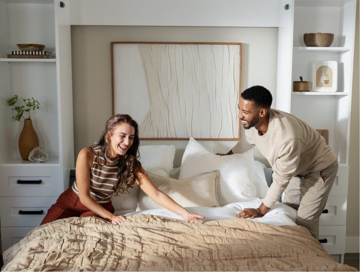 Two people making a Murphy Door® wall bed with cream bedding in a bright, minimalist bedroom with built-in shelving.
