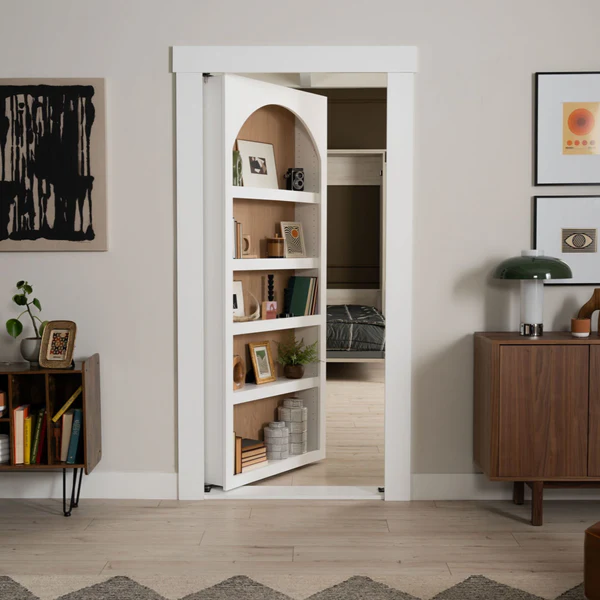 Murphy Door® white bookshelf hidden door, partially open to reveal bedroom, in modern home with wood furniture and art decor.