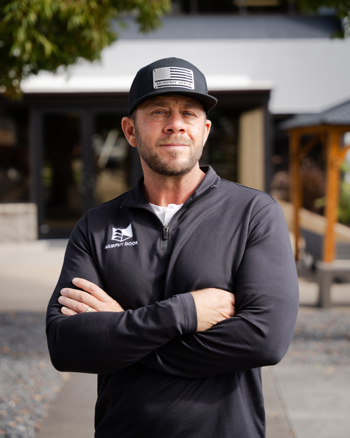 A person wearing a black Murphy Door® zip-up jacket and American flag cap standing with arms crossed outdoors.