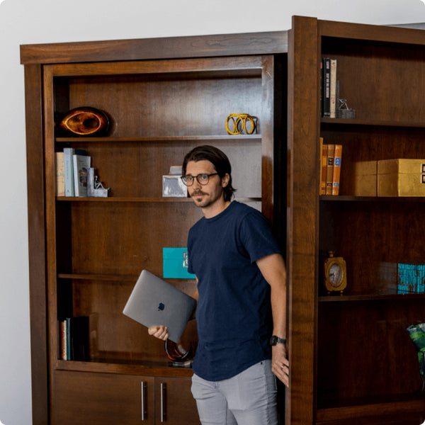 Person standing by Murphy Door® hidden bookcase door in rich wood finish, holding laptop, showcasing seamless storage solution.