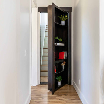 Murphy Door® bookcase hidden door in dark wood, partially open revealing staircase, decorated with plants and books on multiple shelves.