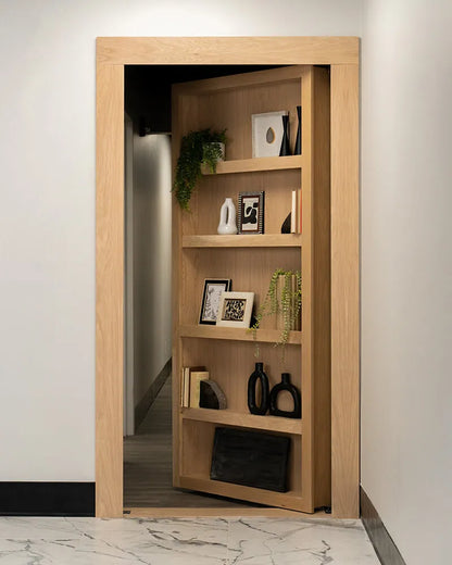 Murphy Door® wooden bookcase hidden door in light oak with decorative plants, frames, and vases on shelves, revealing hallway passage.