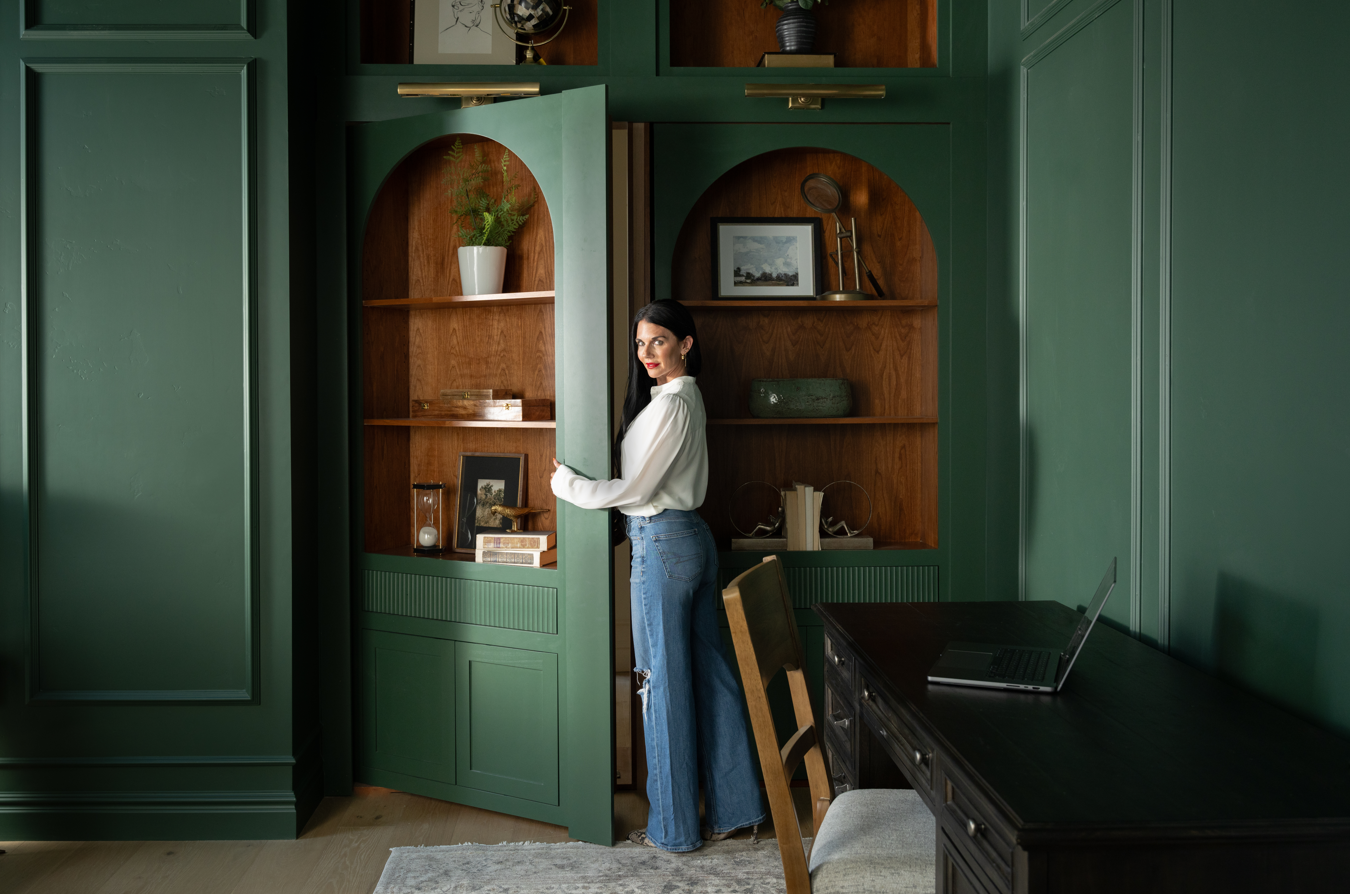 Woman opening Murphy Door® green paneled hidden bookcase door revealing wooden shelving with decorative items