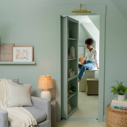 Murphy Door® hidden bookcase door in sage green revealing a nook with person sitting on windowsill, elegant home decor.
