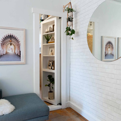 Murphy Door® hidden bookcase door in white, featuring shelves with plants and decor, beside a blue couch and round mirror on brick wall.