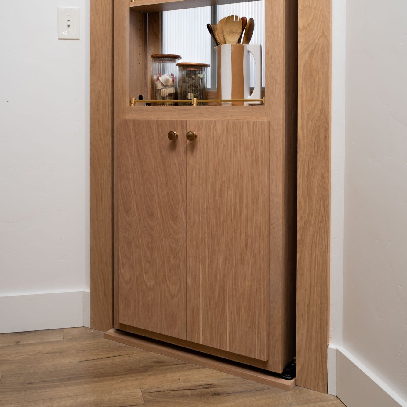 Murphy Door® oak wood hidden cabinet with top shelf displaying kitchen utensils and glass storage jars with wooden lids.