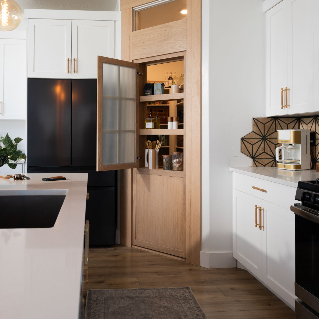 Murphy Door® pantry hidden door in light oak with glass panel, seamlessly integrated into modern white kitchen with black appliances.
