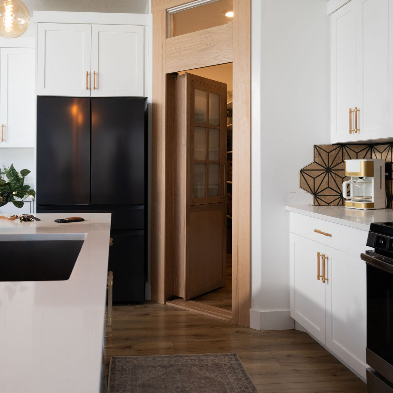 Murphy Door® pantry hidden door in natural wood finish, connecting stylish kitchen with white cabinets, black appliances and geometric backsplash.