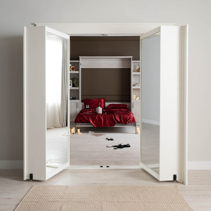 Murphy Door® white bifold doors revealing wall bed with luxurious red bedding, built-in shelving, and romantic candles on light wood flooring.