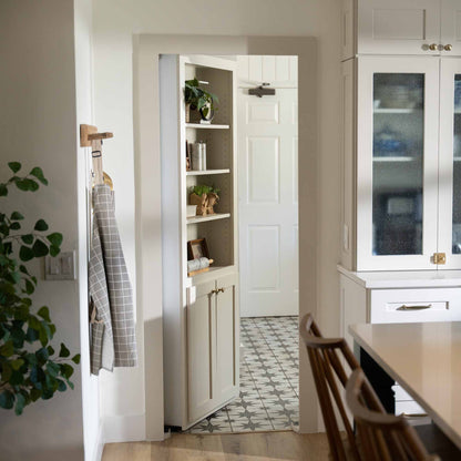 Murphy Door® white bookcase hidden door with shelving, leading to tiled bathroom, in modern kitchen with plants and cabinet storage.