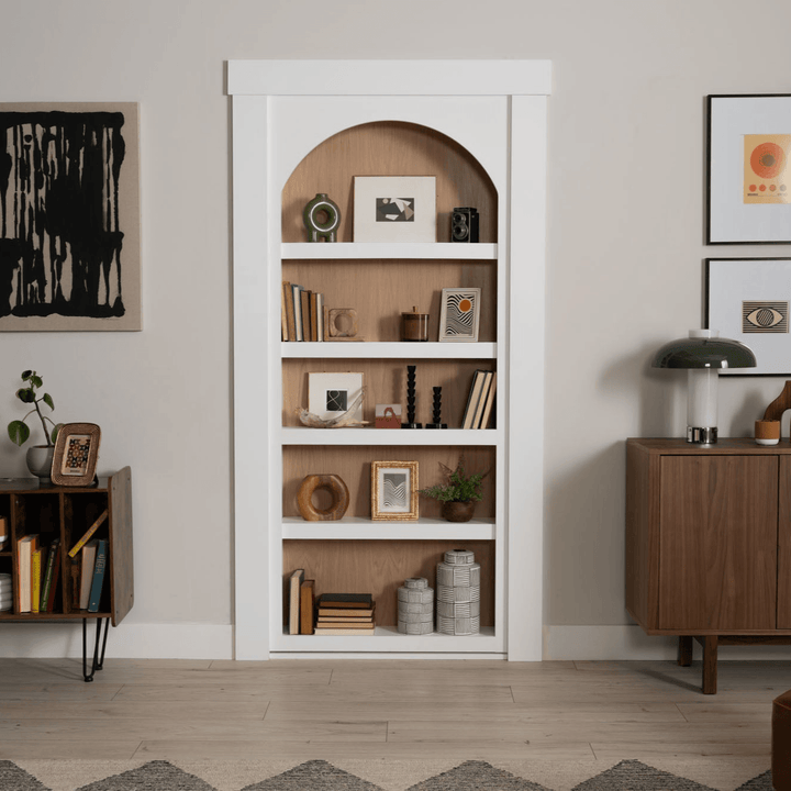 Murphy Door® white bookcase hidden door with arched top, wooden backing, and decorative items displayed on five shelves.