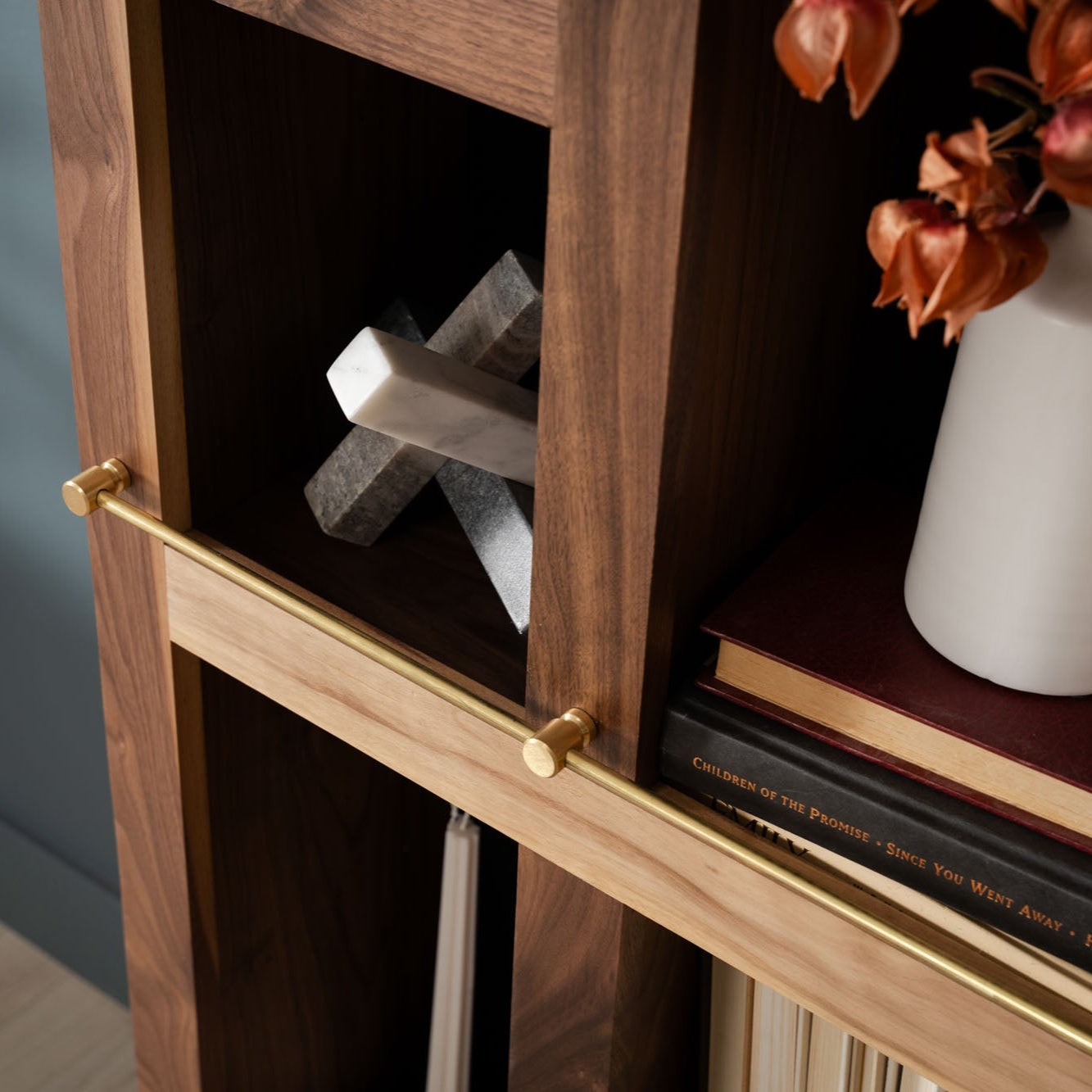 Murphy Door® wooden bookshelf with brass hardware, featuring decorative stone cross, white vase with orange flowers, and stacked books.