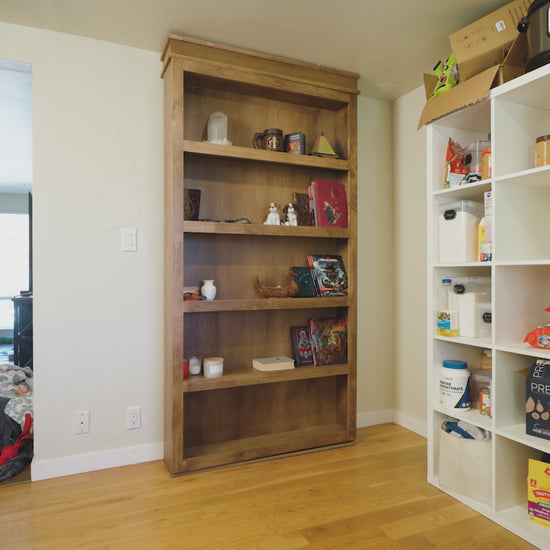 Wooden Murphy Door® bookshelf with decorative items on display, positioned between white pantry shelving and hardwood flooring.