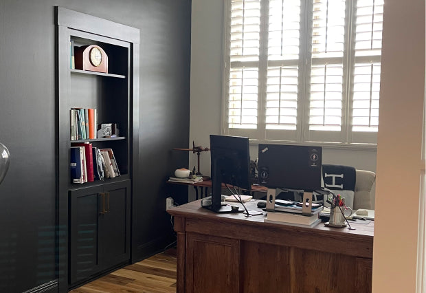 Murphy Door® bookcase hidden door in black, alongside a wooden desk with computer monitors and white shuttered windows.