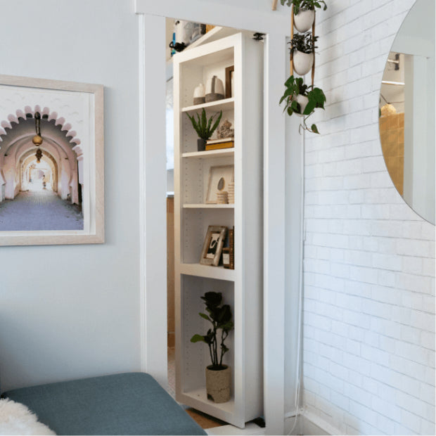 Murphy Door® white bookshelf hidden door displaying plants, decor items and photos, beside white brick wall and round mirror.
