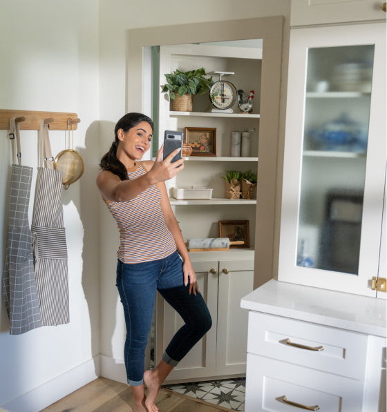 Woman taking selfie in kitchen with Murphy Door® built-in shelving solution featuring decorative items and white cabinetry.